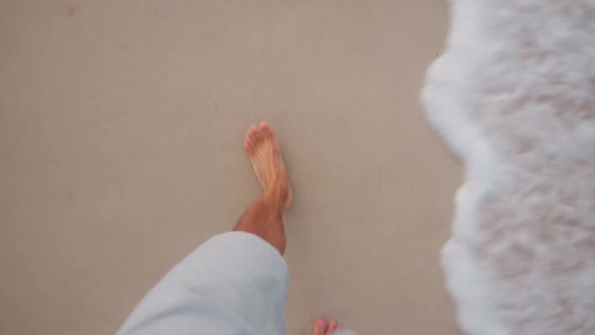 A firstperson perspective shot of a persons bare feet walking through the shallow foamy water of an incoming wave on a sandy beach