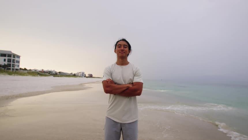 A person stands on a foggy beach with their arms crossed they are wearing a white tshirt and grey shorts looking directly into the camera with a slight smile on their face