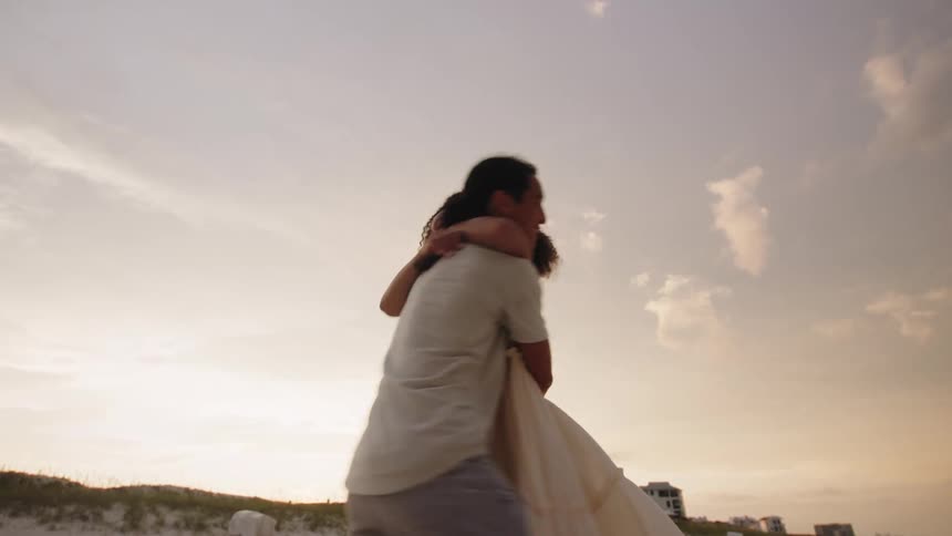 The video shows two people a man and a woman running on a sandy beach at sunset the man has long dark hair tied back in a ponytail and the woman has long curly hair at the end they stop and embrace each other