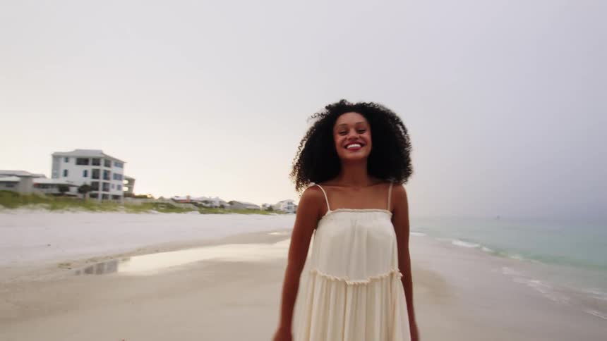 A young woman wearing a white dress stands on the beach sand with long curly hair at midday