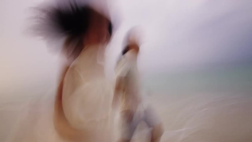 A young adult couple runs barefoot on a sandy beach at the edge of the ocean with the woman wearing a light colored dress and the man wearing shorts and a t shirt creating motion blur in the scene