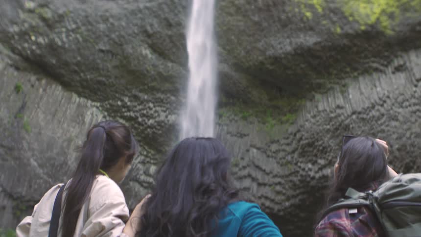 Three friends stand together near a waterfall admiring the view they are looking at the cascading water and the rocks behind it one of the friends with a backpack is pointing at the waterfall