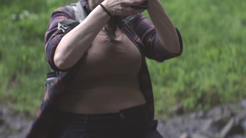 A woman with long dark hair wearing a brown tank top and an unbuttoned red and blue plaid shirt holds a large black professional camera to her eye and films in a green outdoor setting