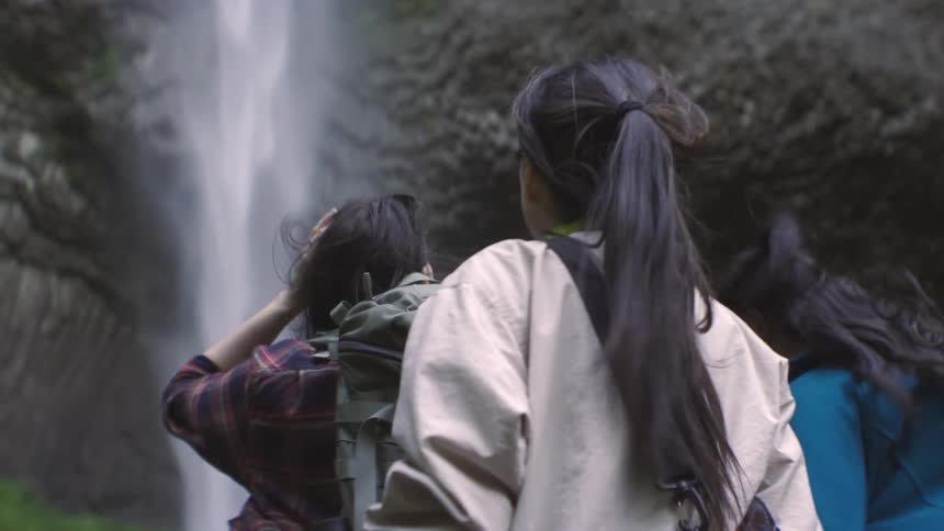 Three females standing on a rock watching a waterfall at midday during a hike carrying backpacks