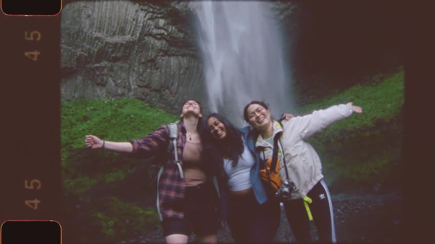 Three smiling women stand with their arms around each other in front of a tall rocky waterfall with green foliage all wearing casual outdoor clothing and backpacks