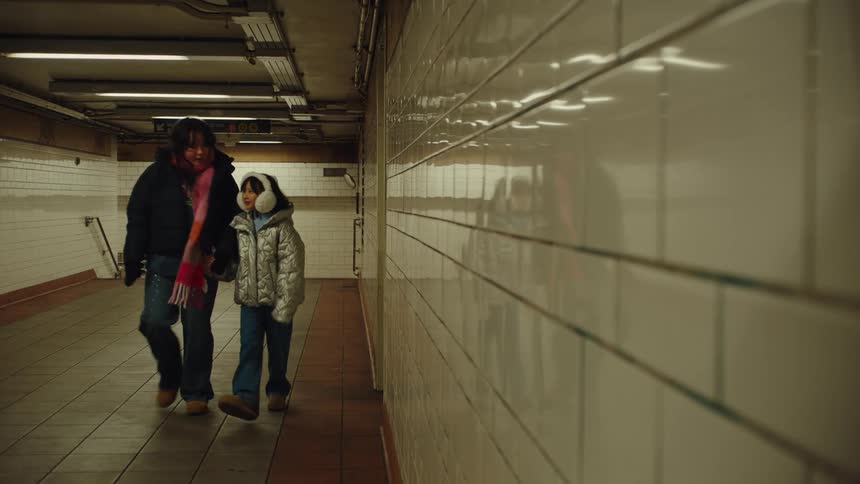 A woman and a young person are walking down an empty subway tunnel the woman has a scarf around her neck and the young person is wearing earmuffs and a shiny jacket they both appear to be bundled up for cold weather