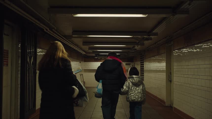 Three people walk through a subway corridor lit by fluorescent lights with walls covered in white tiles