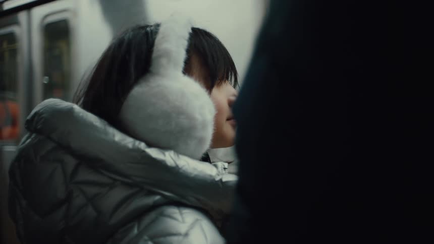 The video shows a young girl with brown hair and white fluffy earmuffs she is wearing a shiny silver puffer jacket and appears to be on a bus or subway