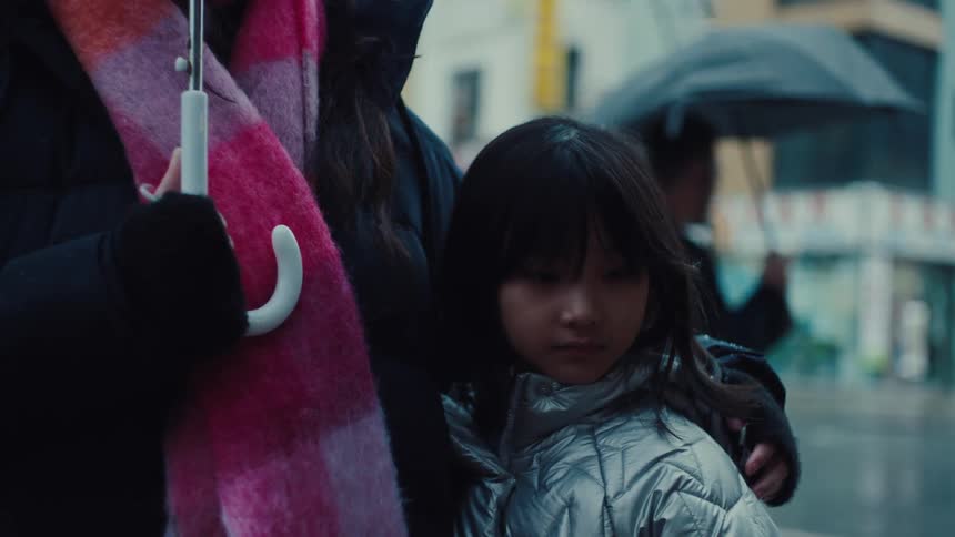 Woman holding a white umbrella while a child in a silver coat leans on her during a rainy day in the city