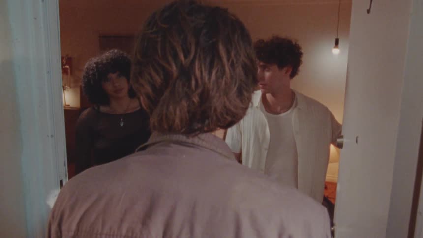 A shot from behind a person shows two friends standing in the doorway of a house the person in the foreground has their back to the camera while the two in the doorway look on with a neutral expression