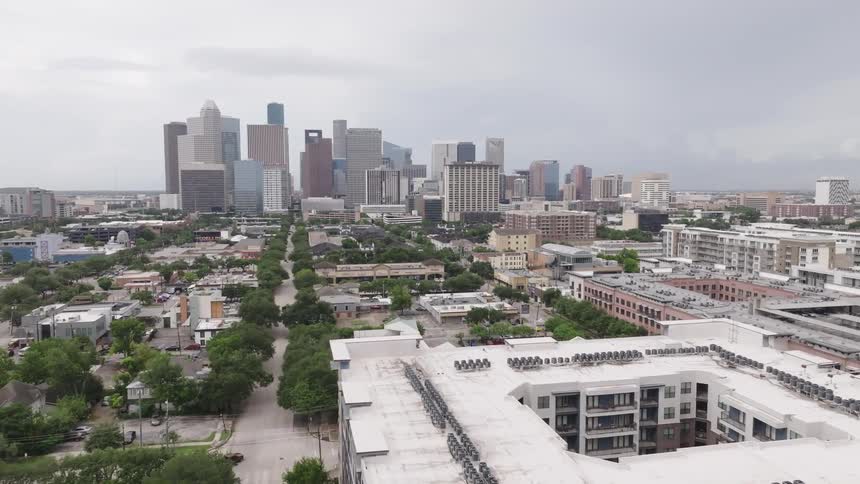 An aerial view shows a large city skyline with many skyscrapers the foreground consists of residential and smaller commercial buildings with a clear street lined with trees and cars