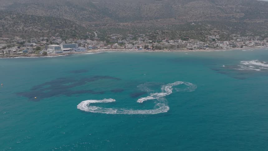A city on the coastline with mountains in the background and a series of boats riding on the ocean water at midday