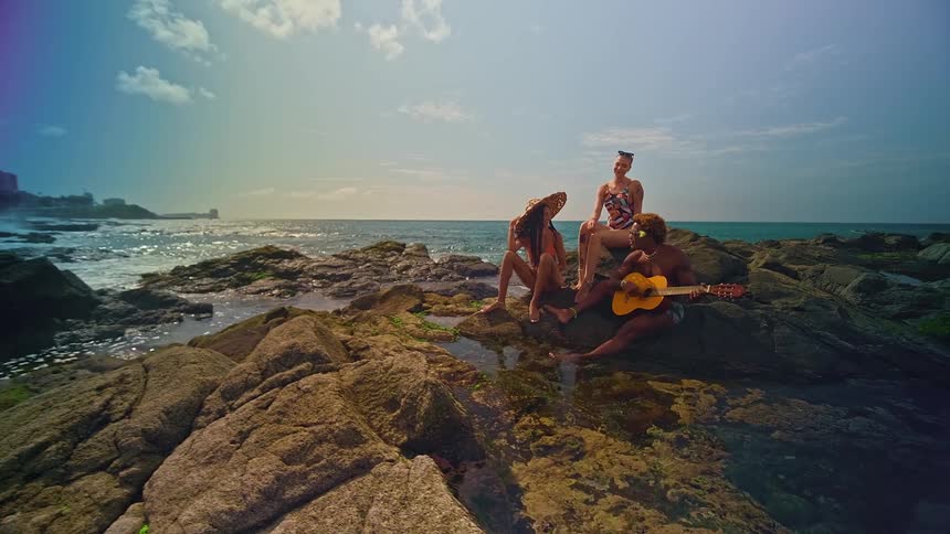 Man sitting on rocks at beach with two women playing guitar