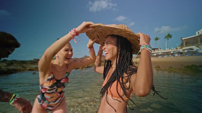 Two women laugh together in shallow beach water as one places a large woven hat on the others head near a lighthouse