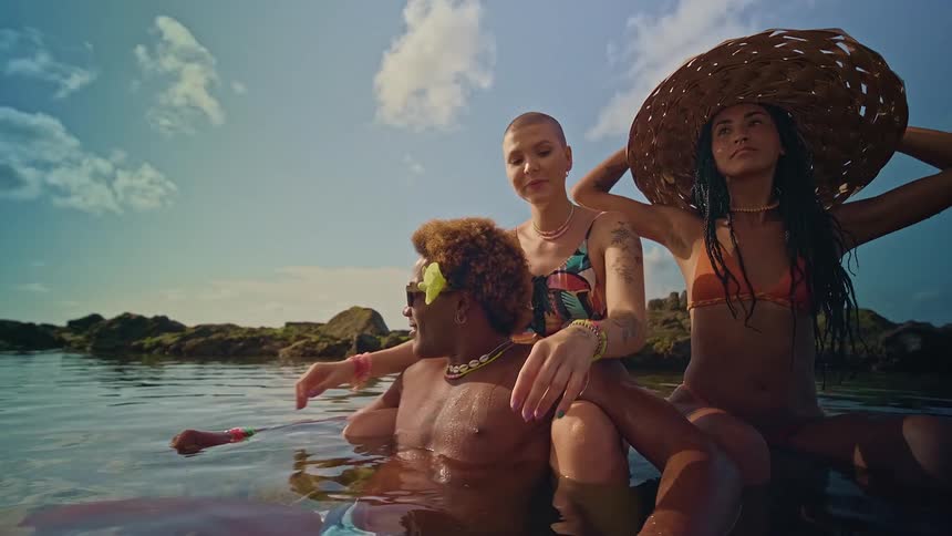 Three people in swimwear enjoy a natural pool by the rocks under a blue sky with some clouds