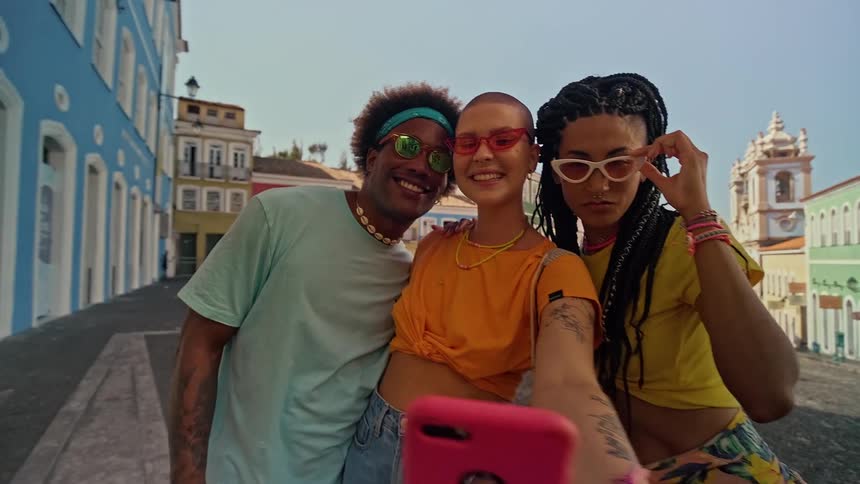 The video shows a group of three friends taking a selfie with a pink phone they are smiling and posing for the camera on a street with colorful buildings in the background