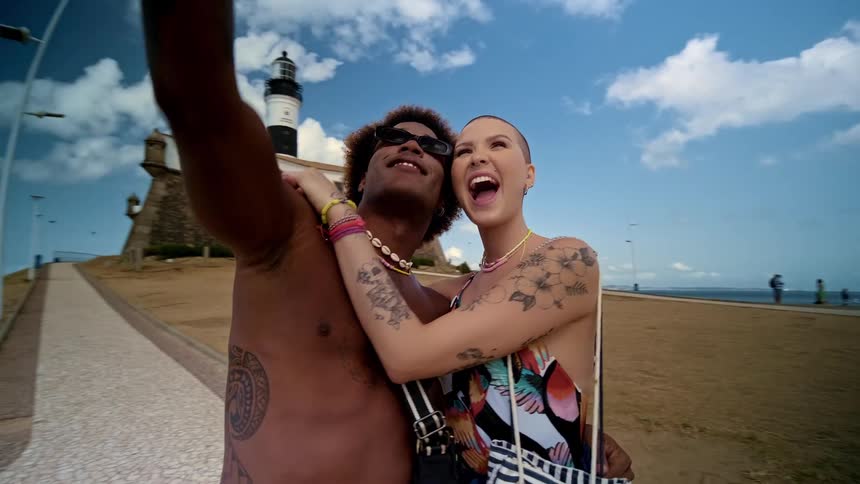The video shows two people a man and a woman taking a selfie together on a sunny beach a lighthouse is visible in the background and the man is smiling widely with a blue bandana and a seashell necklace