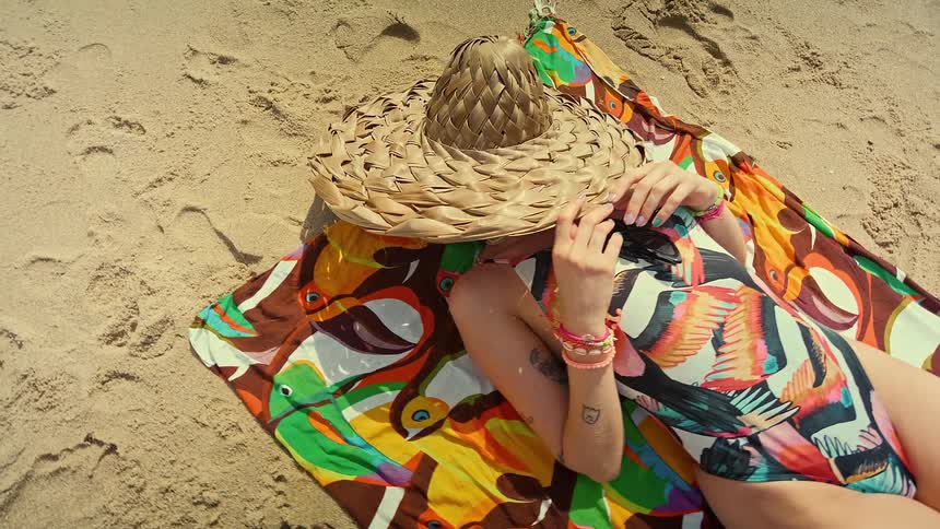 Older woman lying on a towel with a hat on her face on the sand at the beach
