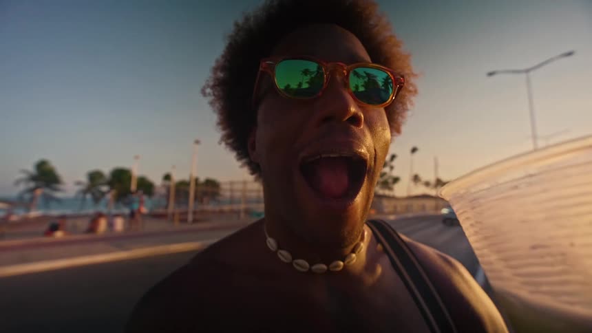 A person with a shell necklace and orange sunglasses smiles and sticks their tongue out at the camera they are holding a clear cup with a drink in it and a beach or boardwalk is visible in the background
