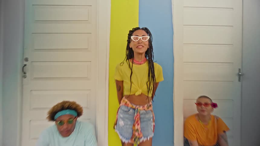 A very short video clip shows three people posing for the camera the person in the middle is standing while the other two are sitting on the ground to either side of them the person in the middle wears a yellow tshirt tied at the waist denim shorts