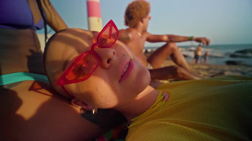 The video shows two people a man and a woman relaxing on a sandy beach the woman is in the foreground with her head resting on someones lap wearing red heartshaped glasses in the background a man with curly hair is sitting on the sand