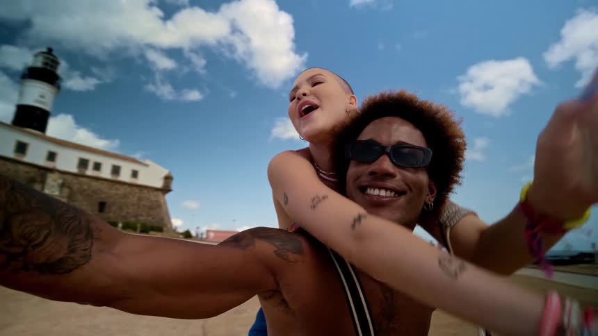 The video shows two people a man and a woman taking a selfie together on a beach a lighthouse is visible in the background the man has afrostyle hair is smiling and wears a blue bandana and a seashell necklace the woman has a shaved head is laugh