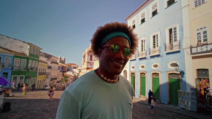A black male in a light blue shirt and seashell necklace walks in a colorful colonialstyle street with pastel buildings and a clear blue sky
