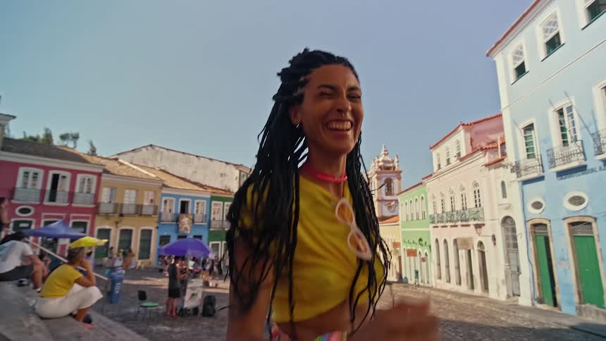 A person with long braided hair and a nose ring is smiling and posing for the camera on a street they are wearing a yellow tshirt tied at the waist and a pair of white sunglasses are hanging from the collar of their shirt