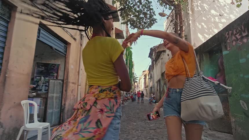 Two joyful women one with long dreadlocks and one with a shaved head walk down a cobblestone street while holding hands and smiling in front of a colorful graffiti covered wall