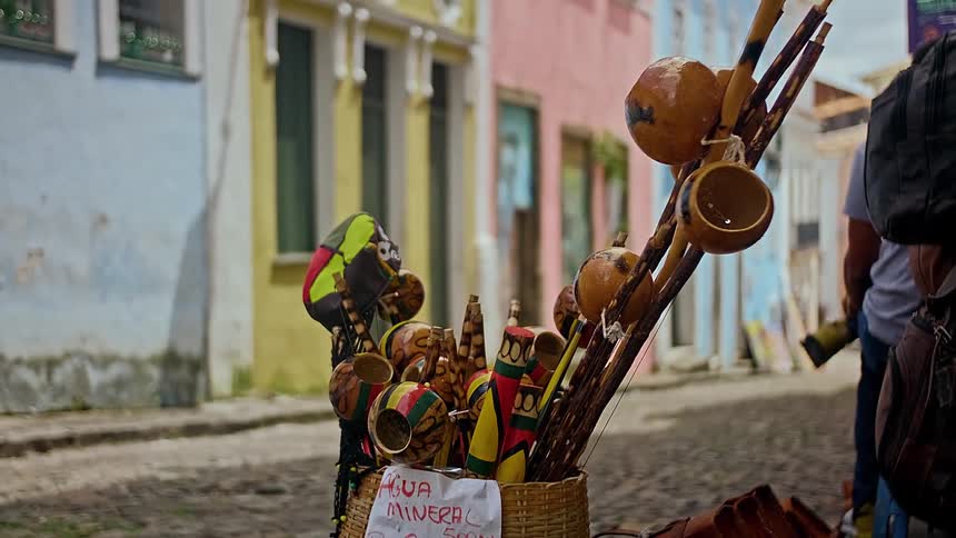 Two baskets containing colorful decorated wooden items and gourd instruments sit on a street next to a price sign for mineral water all in front of a row of light colored buildings
