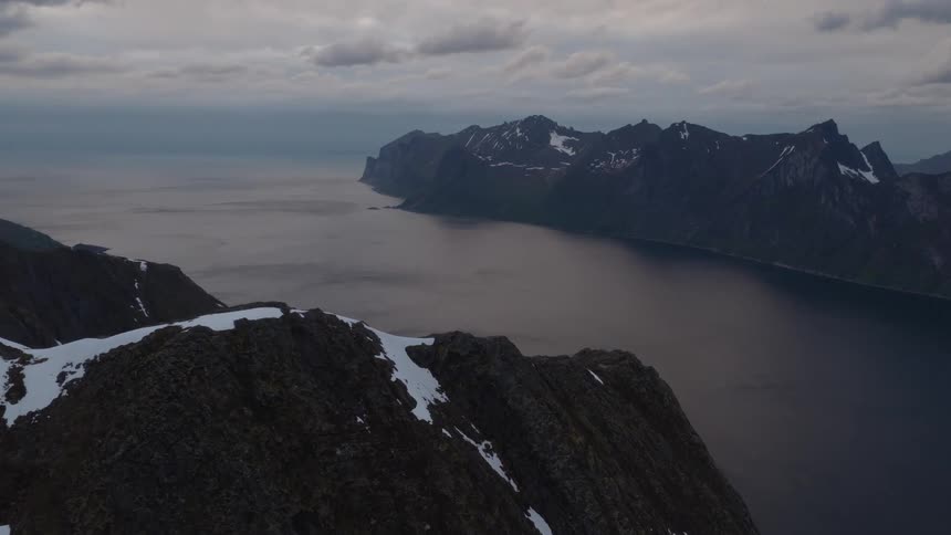 A large dark body of water winds between snow capped mountains under an overcast gray sky
