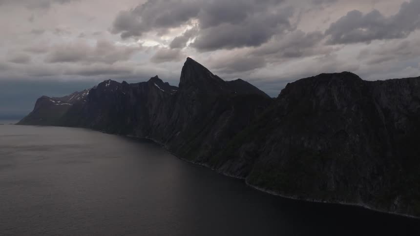 An aerial shot shows a dark rocky mountain range next to a large body of water under a cloudy sky the video slowly zooms out revealing the vast scale of the rugged landscape and the dramatic weather