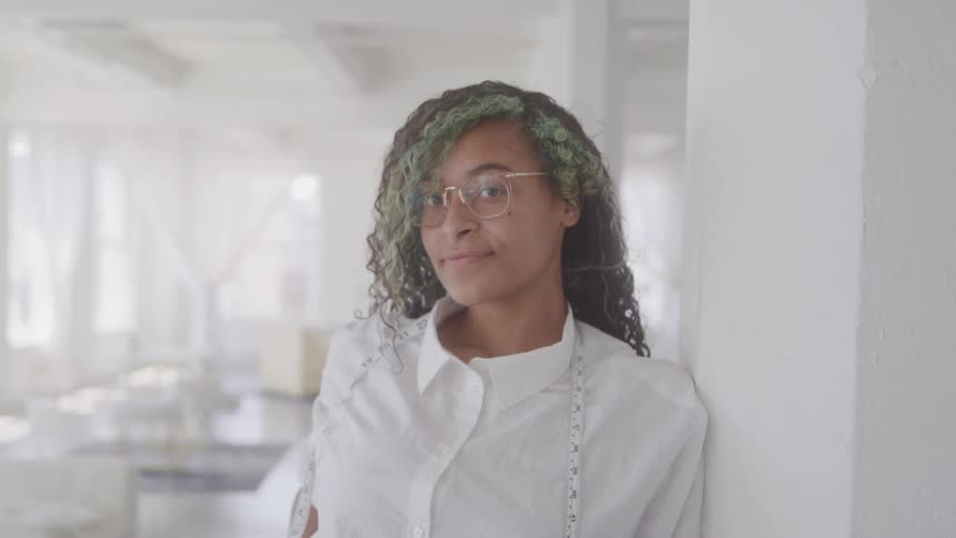 A teenager with green and black hair wearing glasses and a measuring tape around her neck