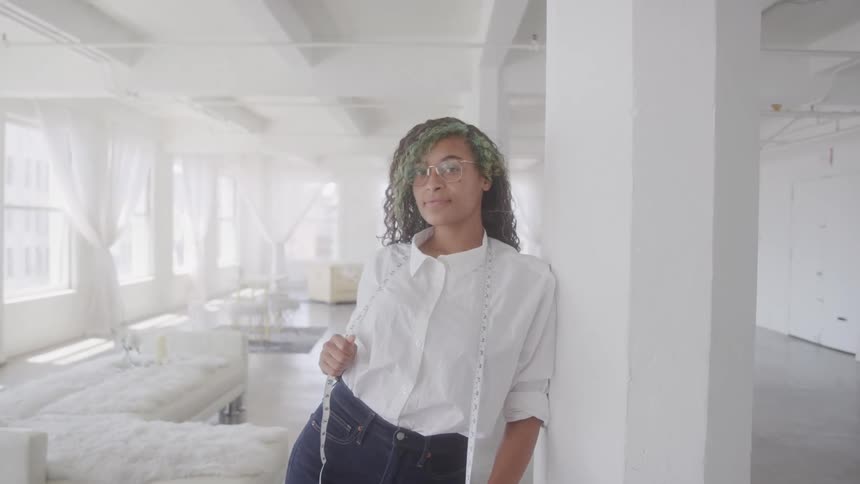 The video shows a woman with curly dark hair and green highlights wearing a white shirt and blue jeans standing in a brightly lit white room with large windows a measuring tape hangs around her neck and she is leaning against a white pillar