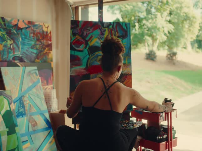 A woman with her hair in a bun is sitting with her back to the camera painting on a large canvas she is surrounded by other colorful paintings in what appears to be a garage or studio space with the outdoors visible behind her