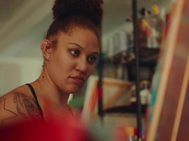 The video shows a closeup of a person with a bun and multiple ear piercings working on a canvas they are painting with a brush and their tattoo is visible on their arm as they move
