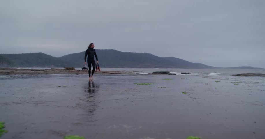 A young man walking on the beach sand wearing a diving suit on a cloudy day