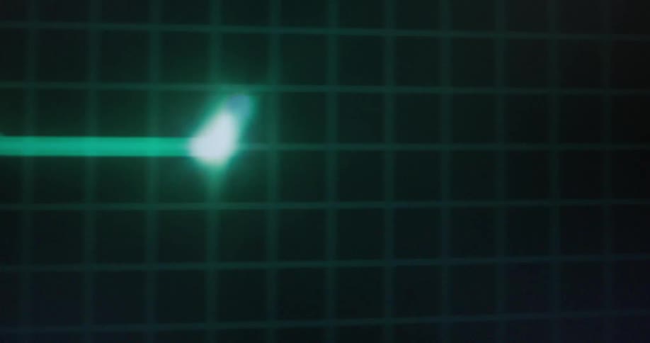A bright green glowing line on a dark grid moves across the screen in a wave pattern the line pulses and moves up and down suggesting a digital waveform or a heartbeat monitor