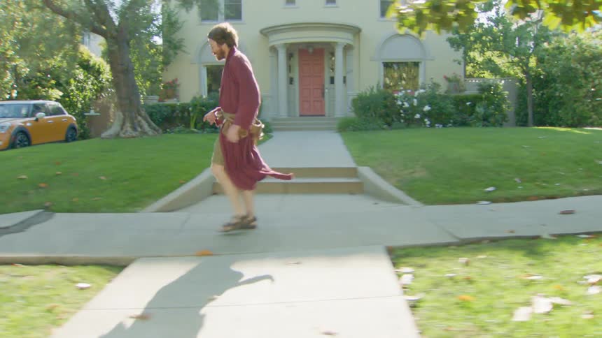 A man in a robe walks quickly barefoot on a sunlit suburban sidewalk passing a manicured lawn and a yellow car