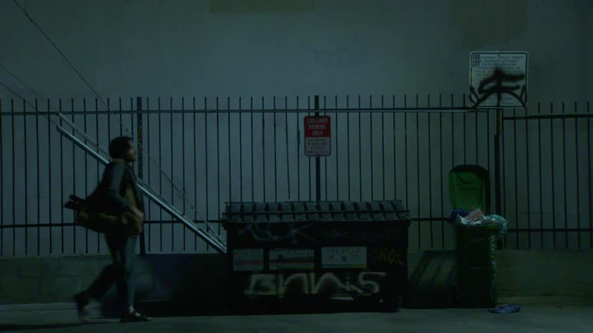 A male walks in dimly a lit alley with a graffitied dumpster and a trash bin against a fenced wall