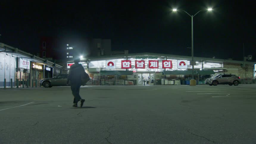 A man with a shoulder bag walks across a parking lot at night in front of a store with a red sign and foreign text all lit by bright streetlights