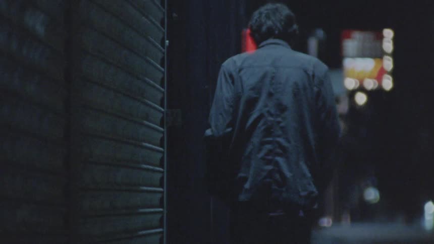 A man wearing a dark jacket walks away from the camera down a dark wet street with blurry lights in the distance