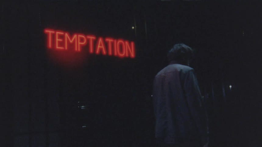 A man walks despondently alone at night illuminated by a glowing red temptation sign