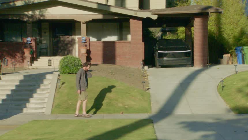 Man in shorts dark jacket and sandals walks on the sidewalk in front of a house with an open garage