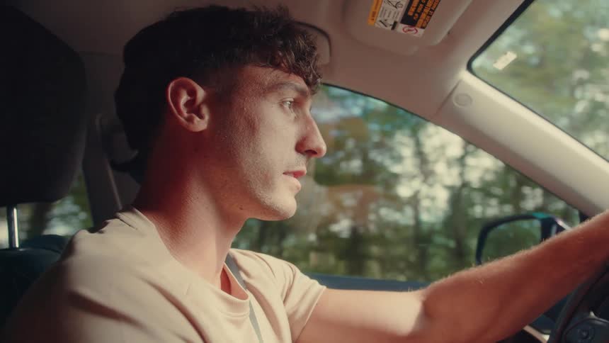 A young man driving a car at midday on a freeway with trees lining the road
