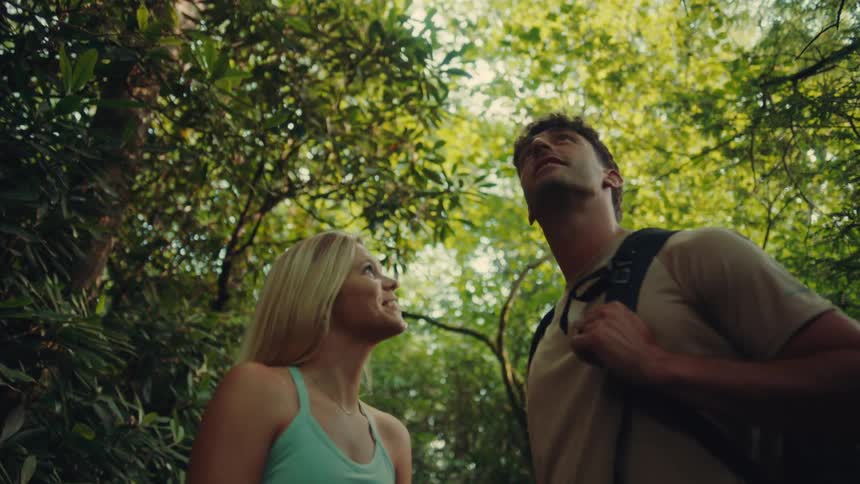The video shows a man and a woman in a forest both looking up the first line is the womans gaze which is directed upward to her right and she is smiling the second line is the mans gaze which is looking straight up and he is wearing a backpack