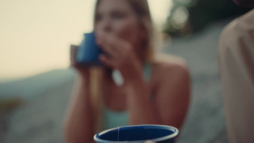 A woman with long blonde hair drinks from a blue mug while sitting on a sunlit hill then smiles with another person holding a matching mug in the foreground