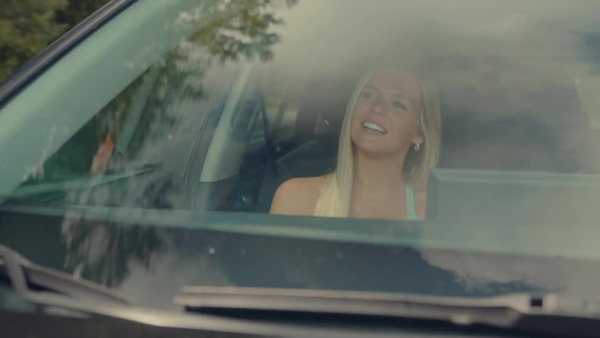 A blonde woman in a light blue tank top is sitting in the drivers seat of a car smiling and looking out of the windshield at one point she points her finger to the right the video is shot from outside the car looking in through the windshield
