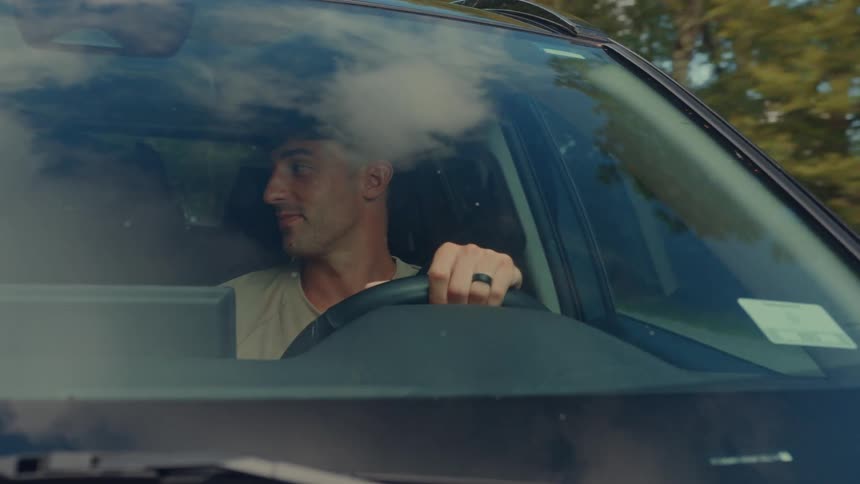 A young man driving a car on a freeway with trees lining both sides of the road at midday