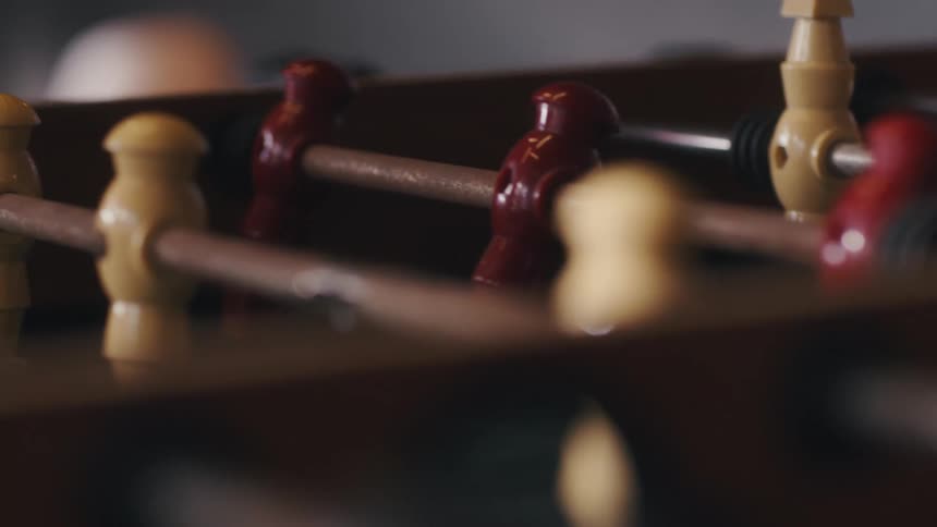 A close view of a foosball table shows rows of light yellow and dark red players on their rods in a dimly lit room